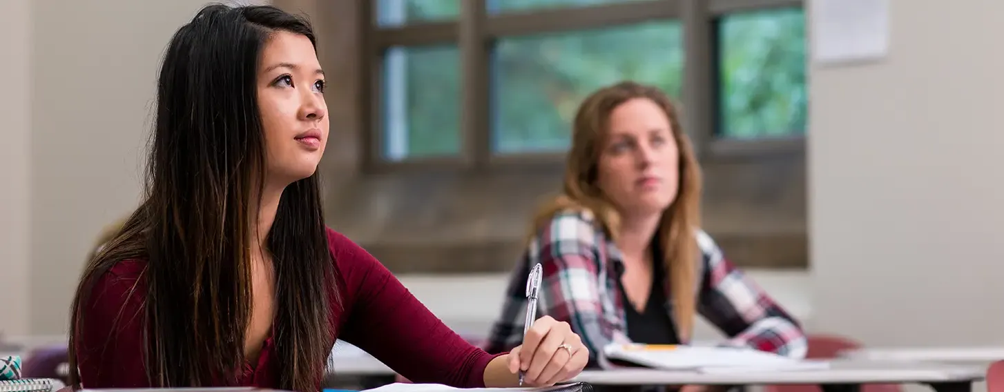 Female students sitting at desks in a classroom with books, taking notes.